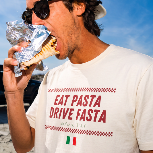 A man wearing a white t-shirt with the text 'EAT PASTA DRIVE FASTA' and a small Italian flag graphic, eating a pasta sandwich on a beach under a blue sky.